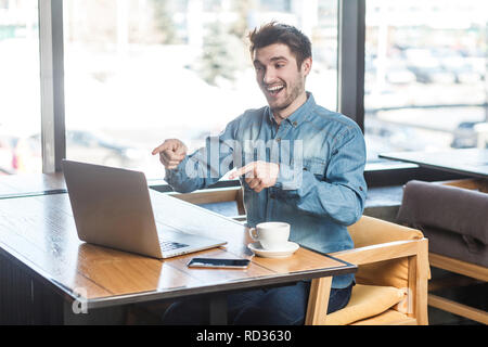 Yes, it`s you! Side view portrait of happy handsome bearded young freelancer in blue jeans shirt are sitting in cafe and making video call on laptop,  Stock Photo